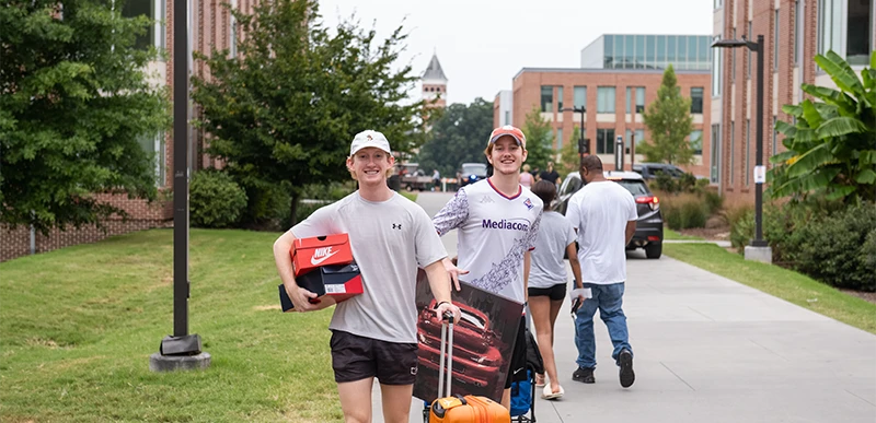 Two male students wearing athletic wear and baseball caps carry suitcases and boxes down a sidewalk on move-in day. 
