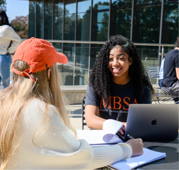 A female student wearing an orange hat and white sweater and a female student wearing a charcoal T-shirt sit together outdoors at a table in front of a laptop and notebooks.