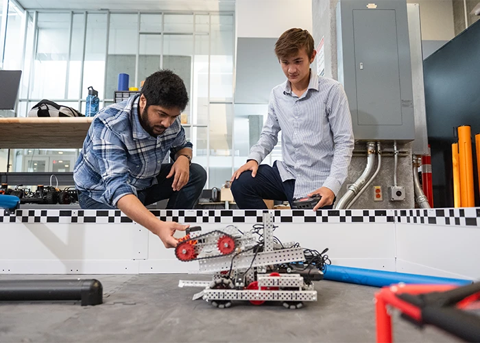 Two male students lean down and inspect a metal robotic device.