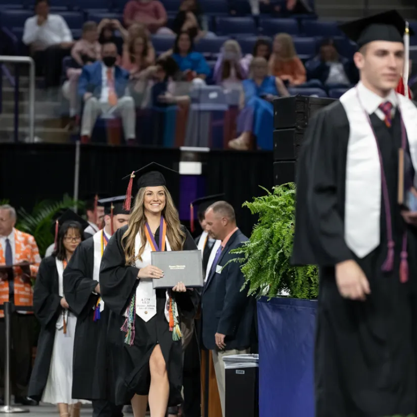 A smiling female college student in a black cap and gown walks across a stage during a graduation ceremony while holding a diploma cover and wearing medals and honors cords, with other graduates visible in the background.