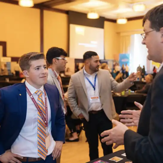 A male student in a navy blue suit and lanyard stands with an older businessman chatting at a networking event.