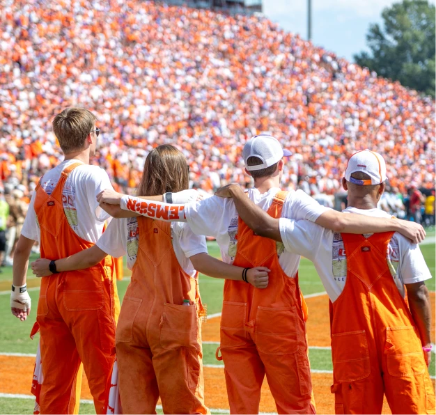 The backs of three male students and a female student who are all wearing orange overalls and white T-shirts with their arms wrapped around each other as they stand on a football field.