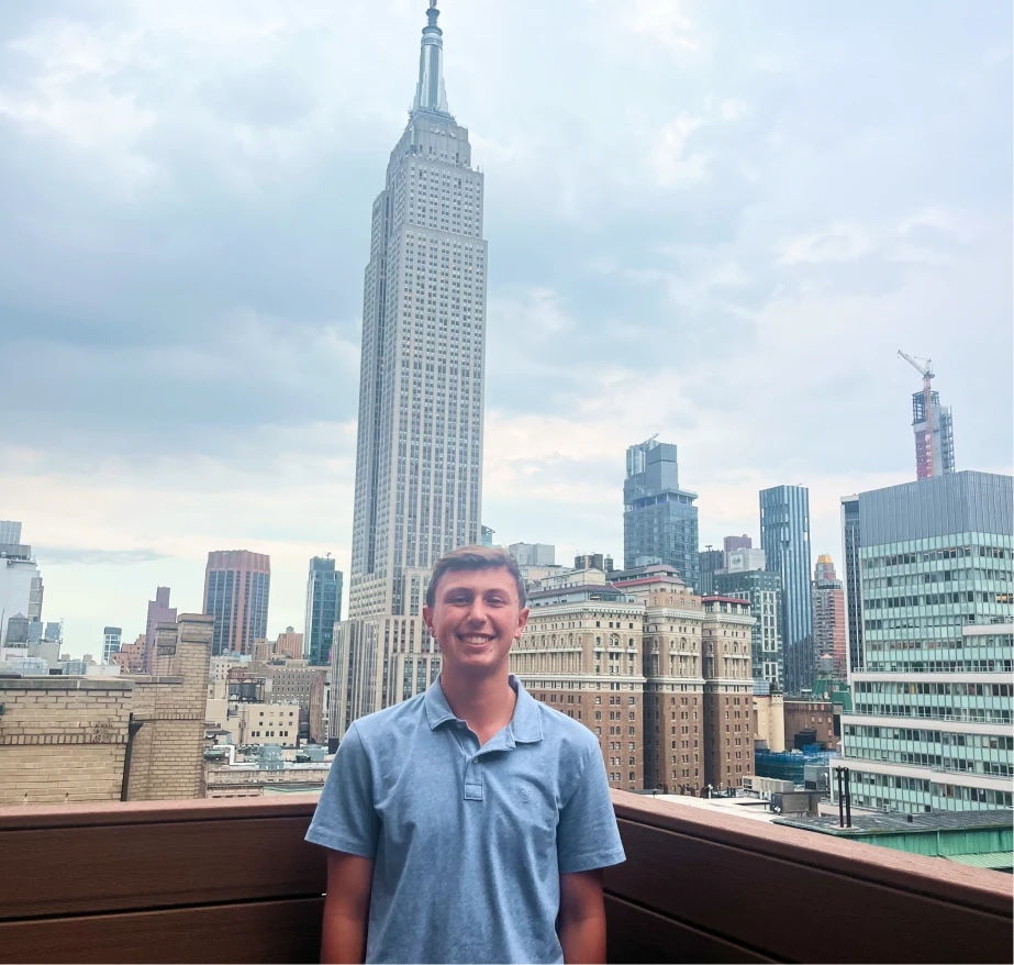 A male student in a light blue polo poses on a rooftop with a busy cityscape in the background.