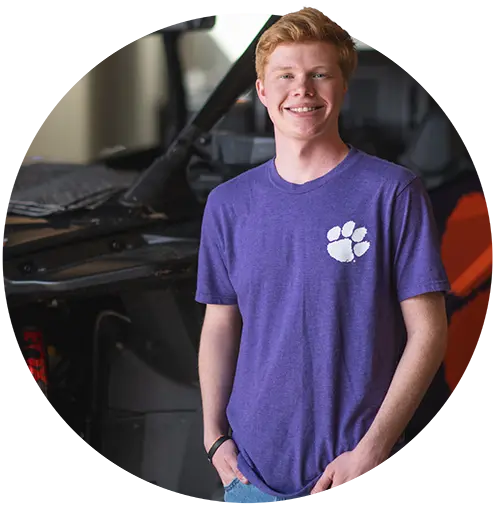 A male student wearing blue jeans and a purple T-shirt with a white Clemson Tiger Paw on it poses in front of the constructed shell of a prototype vehicle.
