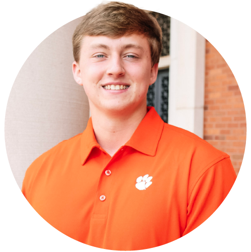 A male student wearing an orange polo with a white Clemson Tiger Paw logo stands next to a concrete column in front of a brick building.
