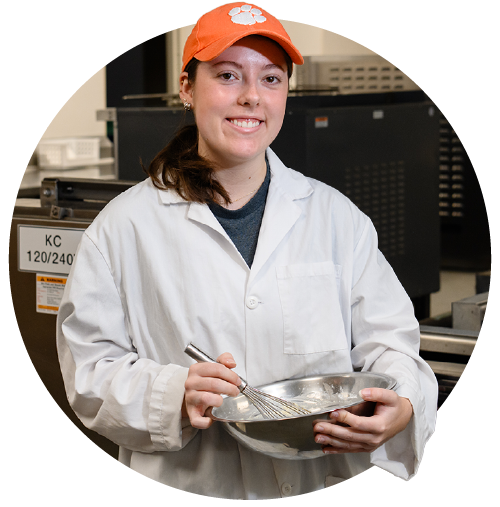 A female student wearing a white chef's coat and orange baseball cap with a white Clemson Tiger Paw logo on it holds a mixing bowl and whisk while standing in a large industrial kitchen laboratory workspace.