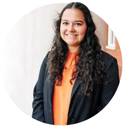 A female student wearing an orange top and black blazer poses in front of an off-white concrete column. 