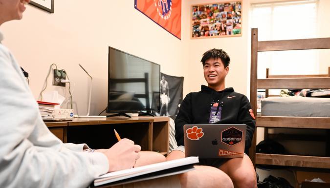 a student sits in his dorm room on a futon holding his laptop and talking to a friend