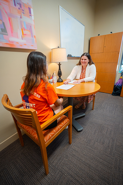 A student and advisor engage in a conversation at a round table in an office setting. The advisor is seated across from the student, who wears an orange shirt, with a lamp and whiteboard visible in the background.