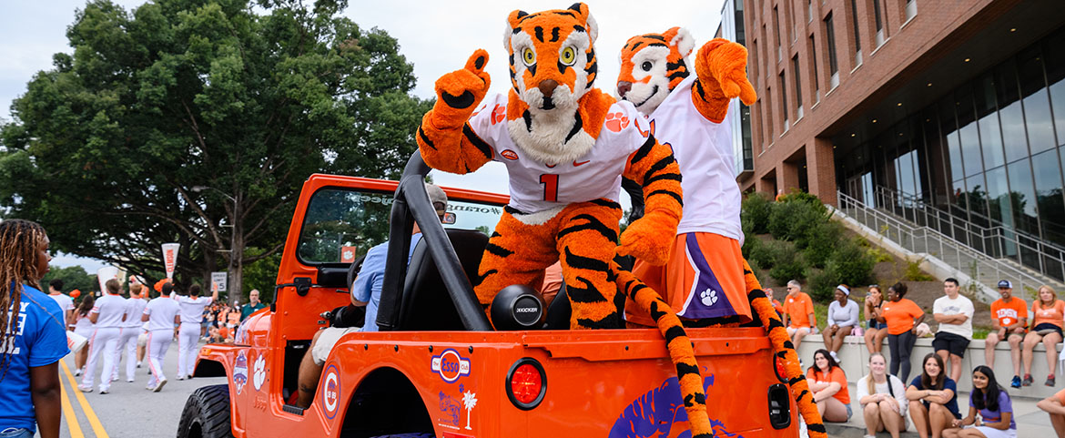 Tiger and Tiger Cub mascots in the back of an orange and purple jeep at the First Friday Parade.