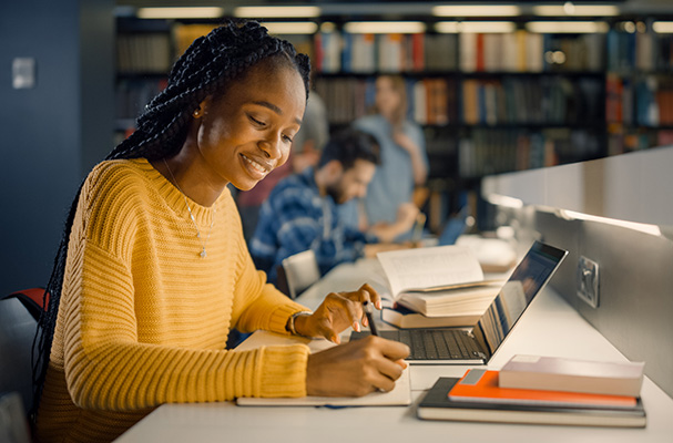 Student studying at a library desk, taking notes beside an open laptop.