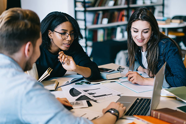Three people collaborating at a table with laptops, notebooks, and printed documents.
