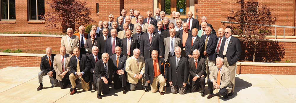 Group photo of class of 1956 alumni gathered outside a building, all wearing formal attire, with some holding canes.