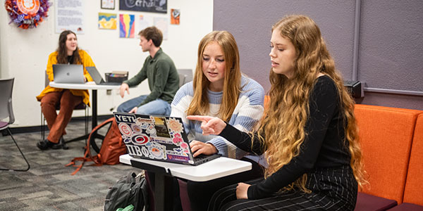 Two students collaborating on a laptop while sitting at a study table in the writing lab, with other students working in the background.