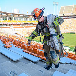 A firefighter in full gear carrying a folded up hose up the stairs in the Clemson football stadium.