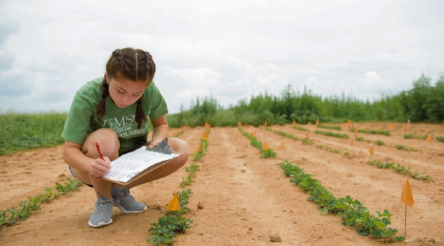 A female student writes in a notepad while crouching beside a row of plants in a field.