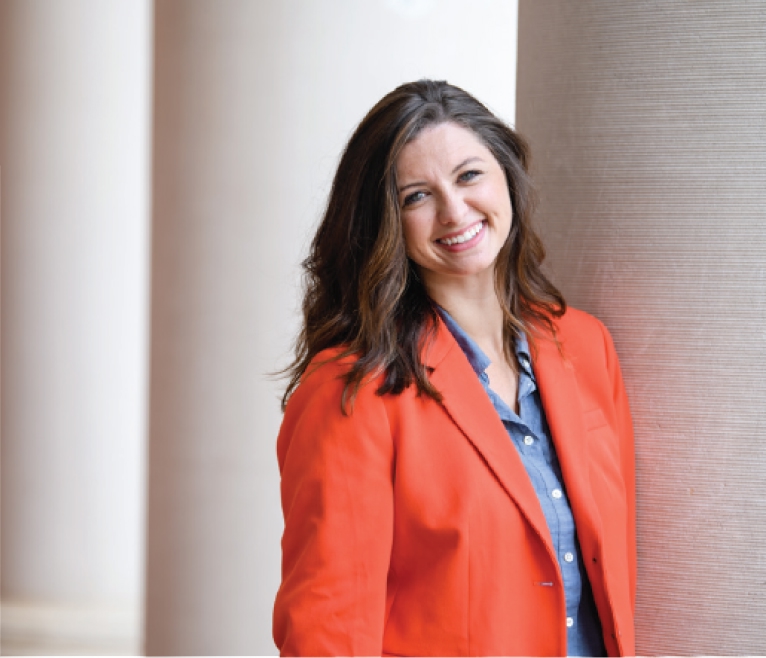 Portrait of a smiling woman wearing an orange blazer, leaning on a column in front of Sikes Hall.