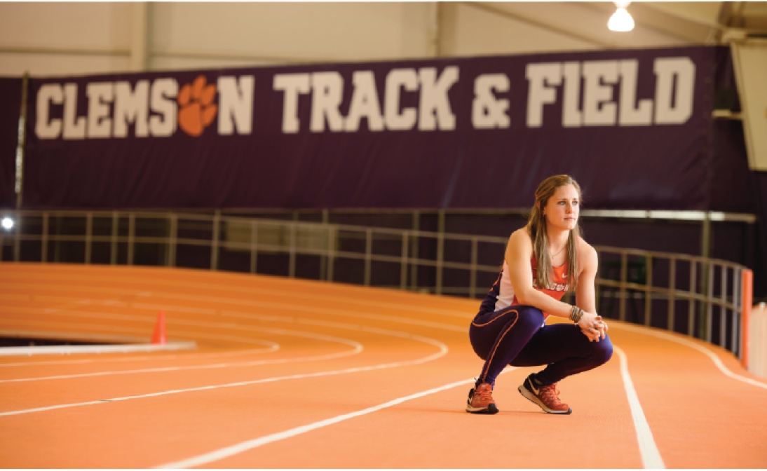 Portrait of a female athlete in orange and purple track uniform crouching on the track with a large 'Clemson track & field' sign behind her.
