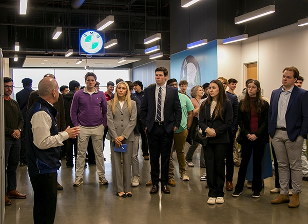 Group of attendees in business attire listening to a speaker in a modern showroom or lobby, with a BMW logo visible overhead.