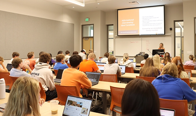 University classroom lecture with students seated at long tables using laptops while an instructor presents slides on a screen at the front of the room.