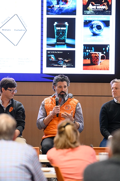 Panel discussion in a classroom, with a man in an orange vest speaking into a microphone while slides of product images are projected behind him.