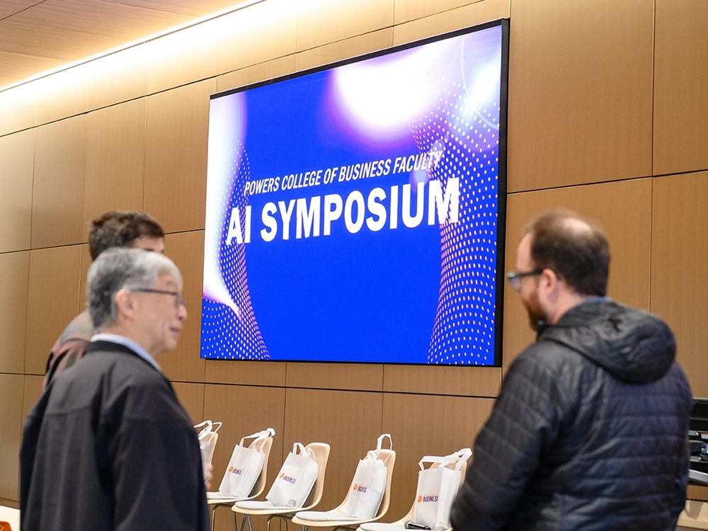 Attendees chat in a lecture hall beneath a large screen displaying “AI Symposium.”.