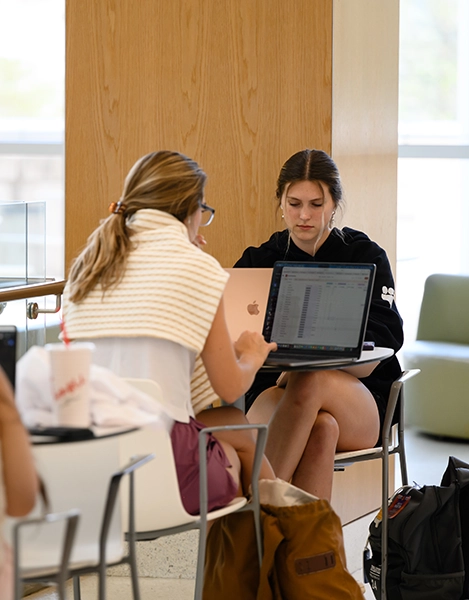 Two students sit at a table working on a laptop in a bright indoor campus lounge, with backpacks and a drink nearby.