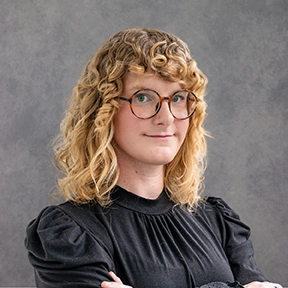 Portrait of a woman with curly hair and glasses, arms crossed, with a gray background.