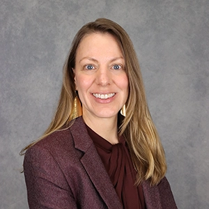 Professional headshot of a woman with long hair, wearing a burgundy blazer and statement earrings, set against a gray background.