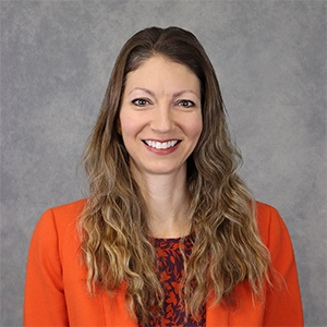 Professional portrait of a woman with long wavy hair wearing an orange blazer and a patterned top against a gray backdrop.