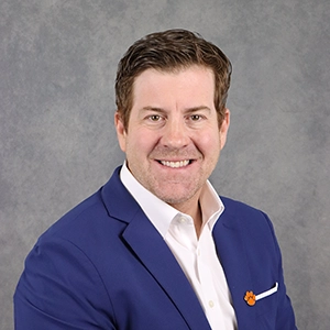 Professional headshot of a man in a blue blazer and white shirt, smiling against a gray backdrop.