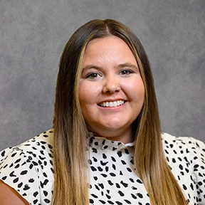 Smiling woman with long straight hair wearing a white patterned blouse against a gray studio background.