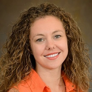 Professional headshot of a woman with curly hair, wearing an orange blouse and smiling warmly.