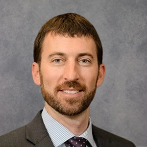 Head-and-shoulders professional portrait of a man in a suit and tie against a gray studio background.