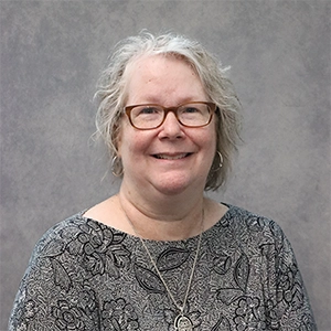 Professional headshot of a woman with glasses, wearing a patterned top, smiling against a gray background.