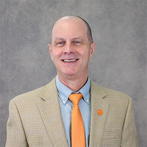 Headshot of a smiling man in a tan blazer and orange tie against a gray background.