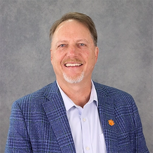 Professional portrait of a smiling man in a blue plaid blazer, with a light blue shirt and an orange emblem on the lapel.