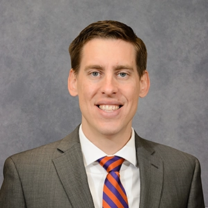Head-and-shoulders professional portrait of a smiling man in a gray suit with a blue-and-orange striped tie against a neutral gray background.