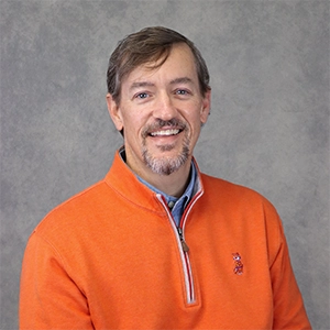 Portrait of a smiling man in an orange sweater with a gray background.