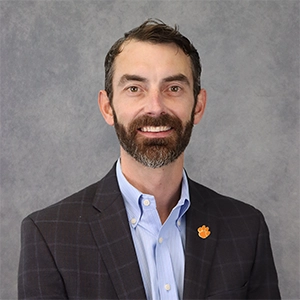 Portrait of a man with a beard wearing a dark blazer and light blue shirt, smiling against a gray background.