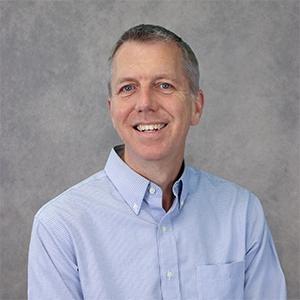 Professional headshot of a smiling man in a light blue button-up shirt against a gray background.