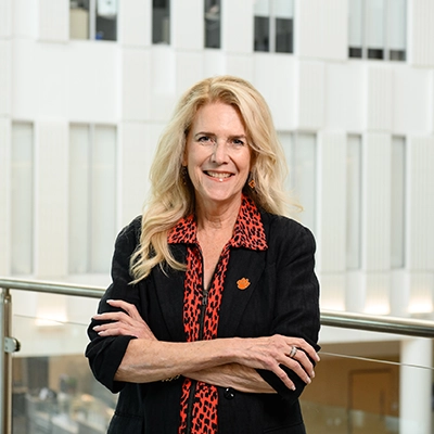 Smiling woman with long blonde hair stands with arms crossed in a modern office atrium, wearing a black blazer and a red patterned blouse.