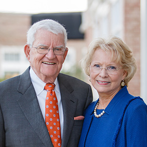 A warm portrait of a smiling elderly couple, both dressed elegantly, with the man in a suit and the woman in a blue outfit.