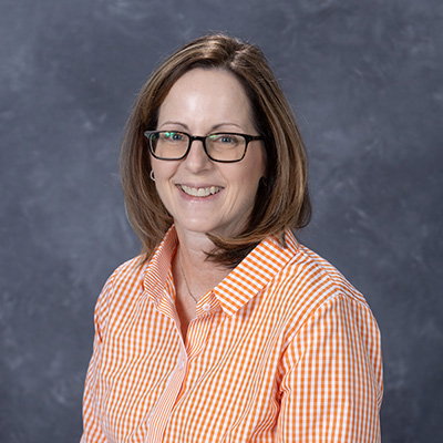 Professional headshot of a smiling woman with glasses, wearing an orange and white checked shirt.