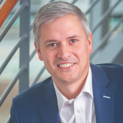 Smiling man in a blue suit and white shirt, shown in a close-up portrait with a modern staircase in the background.
