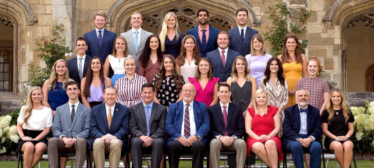 Group portrait of a diverse cohort of individuals, dressed in formal attire, seated in front of a historical building with greenery.