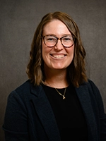 Studio headshot of a smiling person with shoulder-length hair and glasses, wearing a dark blazer over a black top against a neutral background.