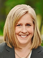 Professional headshot of a smiling woman with shoulder-length blonde hair, wearing a dark blazer and a necklace.