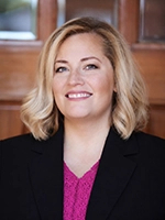 Portrait of a smiling woman with shoulder-length blonde hair wearing a dark blazer over a bright pink top, standing indoors against a warm wood background.