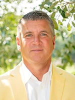 Portrait of a man wearing a pale yellow blazer and white shirt, standing outdoors with leafy greenery in the background.